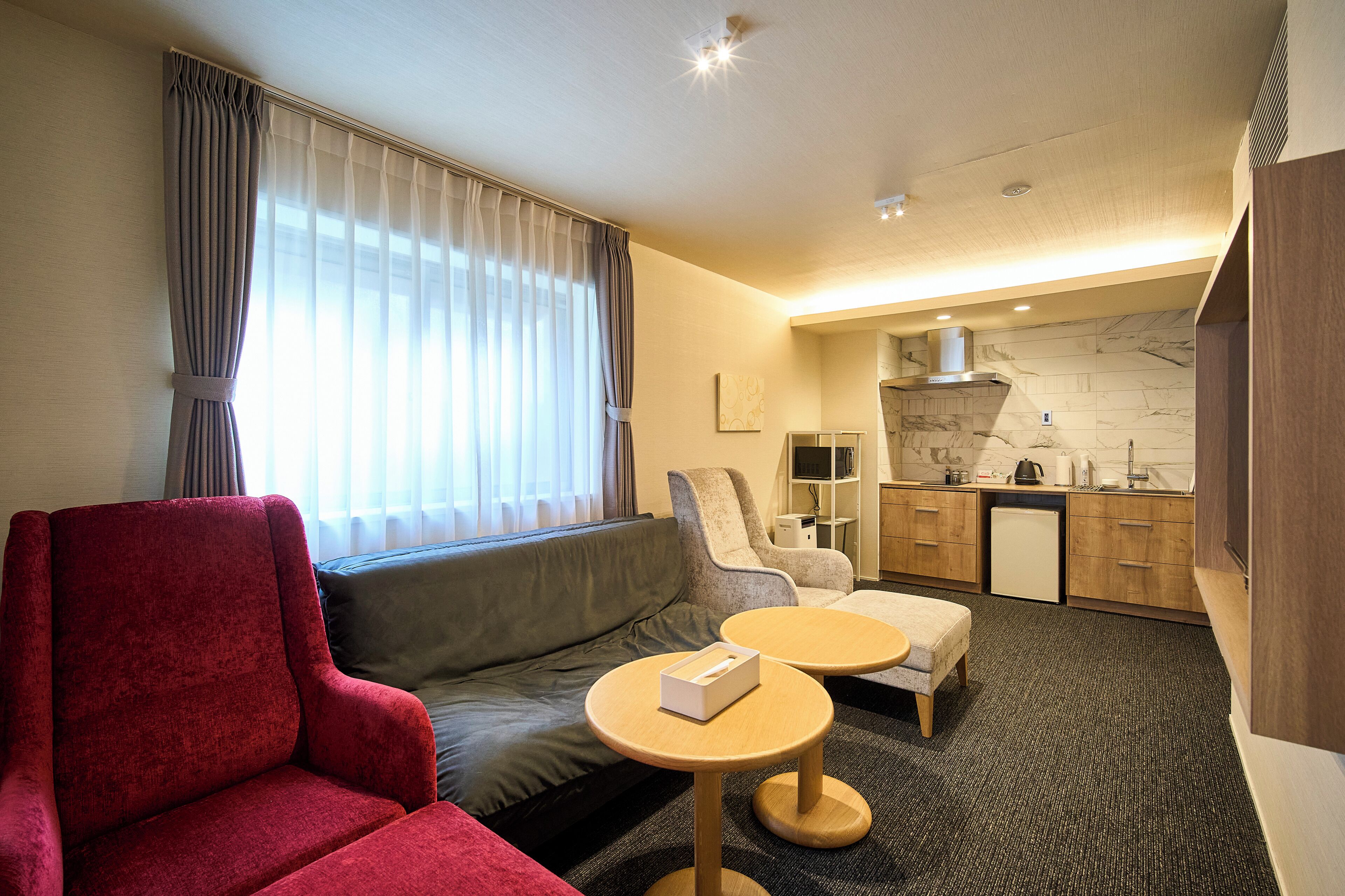 Quarto de hotel moderno com poltrona vermelha, sofá cinzento, mesa de madeira pequena e uma kitchenette ao fundo.