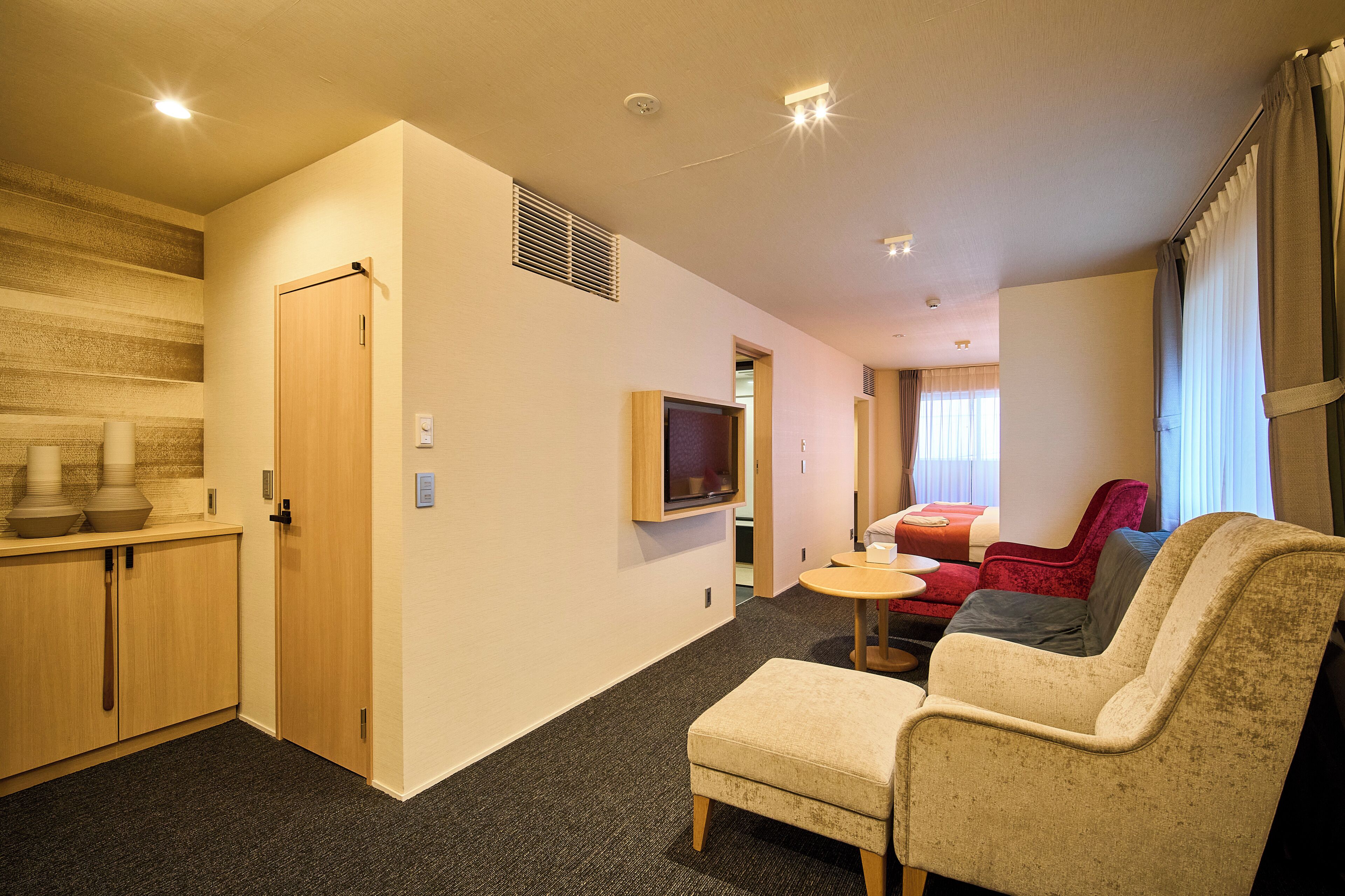 Ein kompaktes Hotelzimmer mit Küchenzeile, einer Sitzecke mit Sofa und Sesseln, einem an der Wand montierten Fernseher und einem Fenster mit Vorhängen.