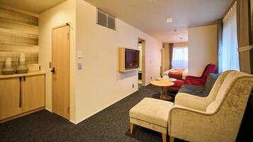 Ein kompaktes Hotelzimmer mit Küchenzeile, einer Sitzecke mit Sofa und Sesseln, einem an der Wand montierten Fernseher und einem Fenster mit Vorhängen.