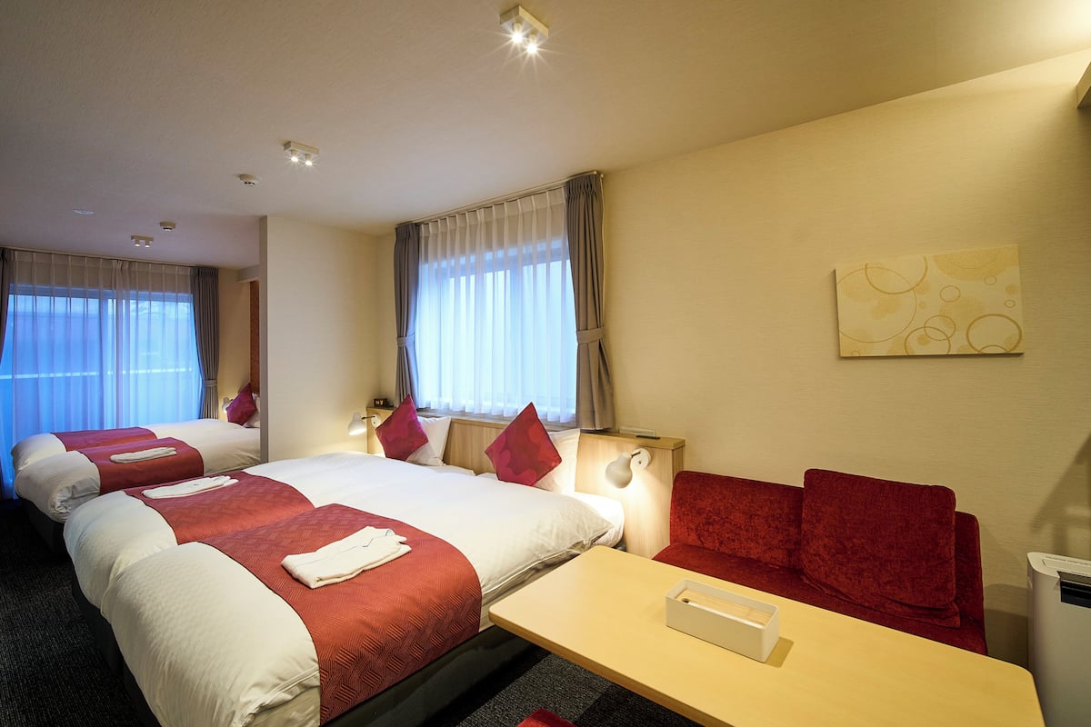 A hotel room with two beds, a sofa, a desk, and a wall-mounted lamp.