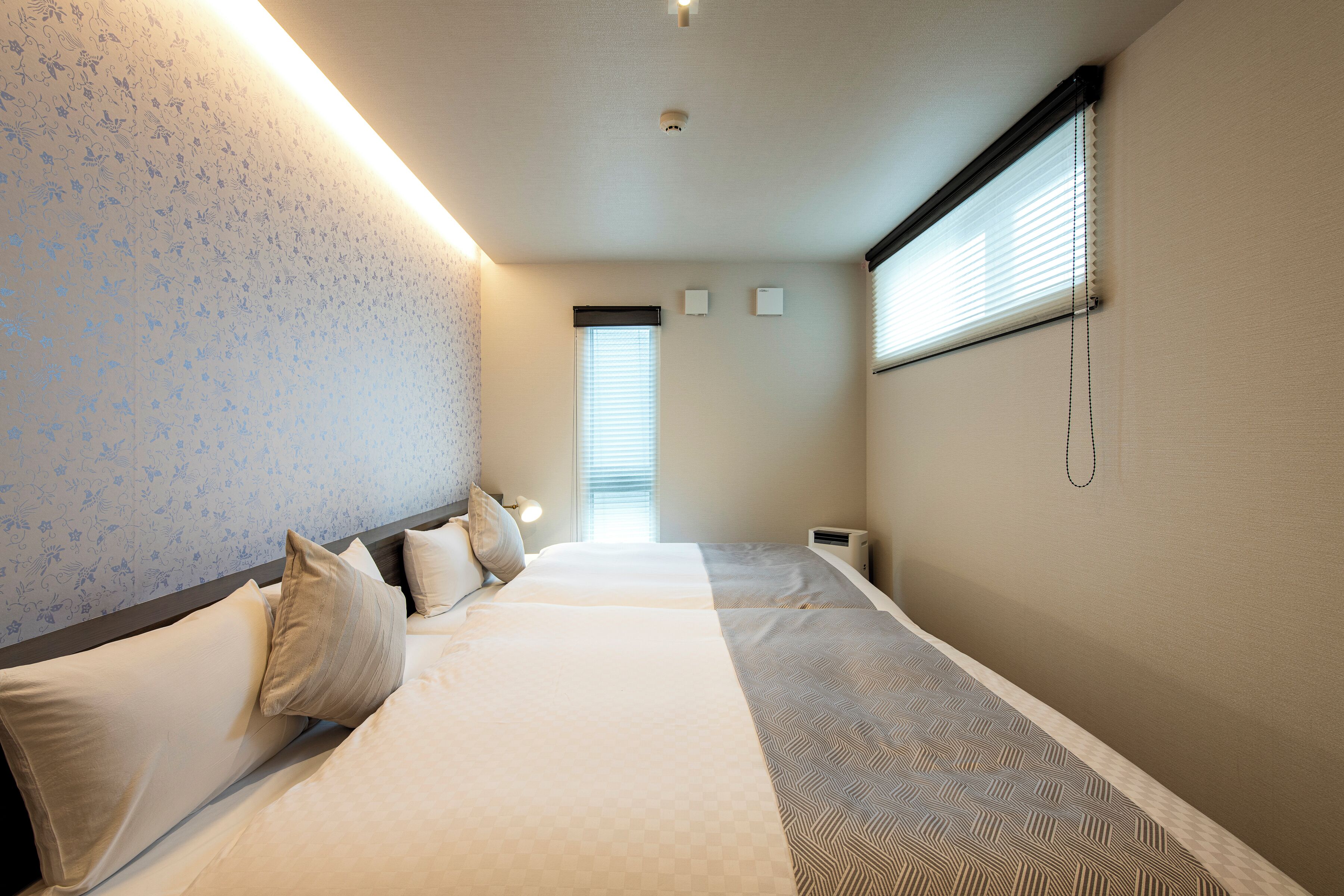 A hotel room with two beds, a window with blinds, a wall-mounted air conditioner, and a ceiling light.