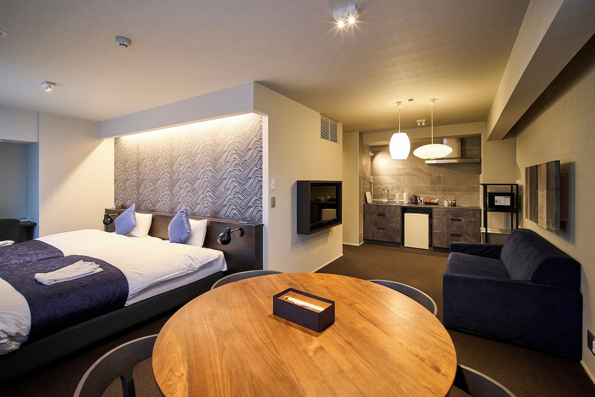 A modern hotel room with a bed, a round wooden table, a sofa, a TV, and a kitchenette.