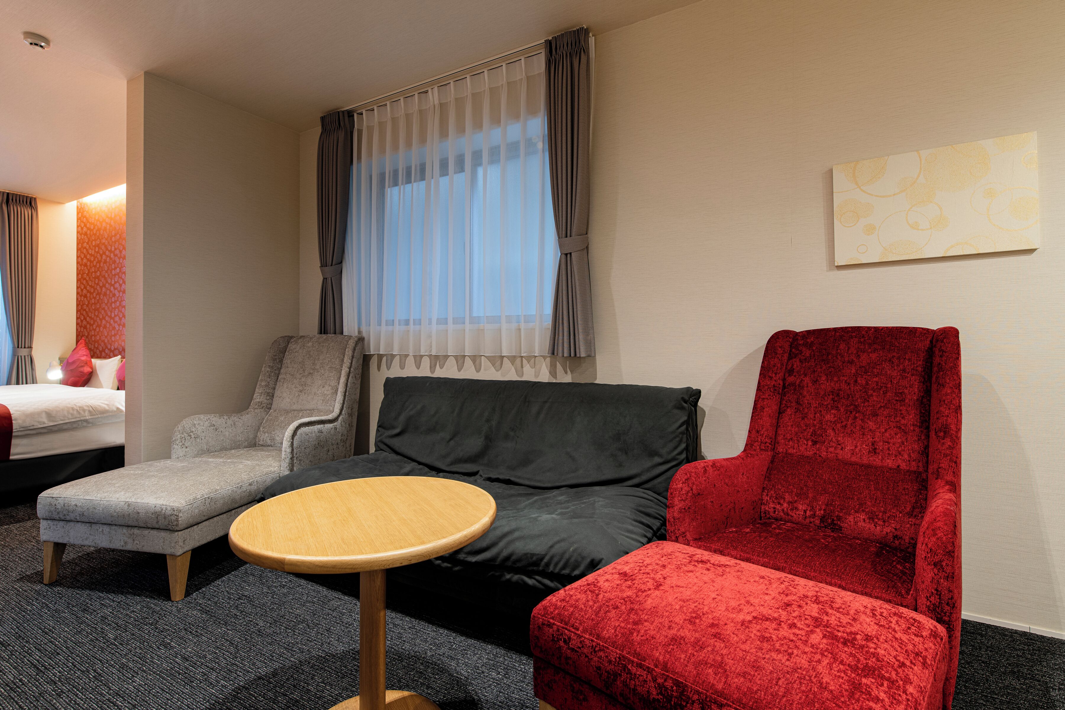 A hotel room with a grey armchair, a red armchair, a black sofa, and a small wooden table.