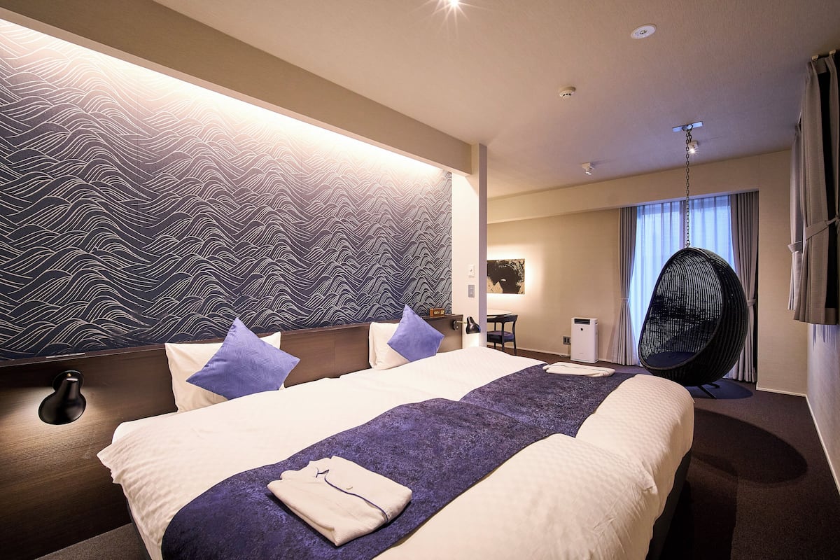 A hotel room with a large bed, a wall with a wavy pattern, a hanging chair, and a small table with a lamp.