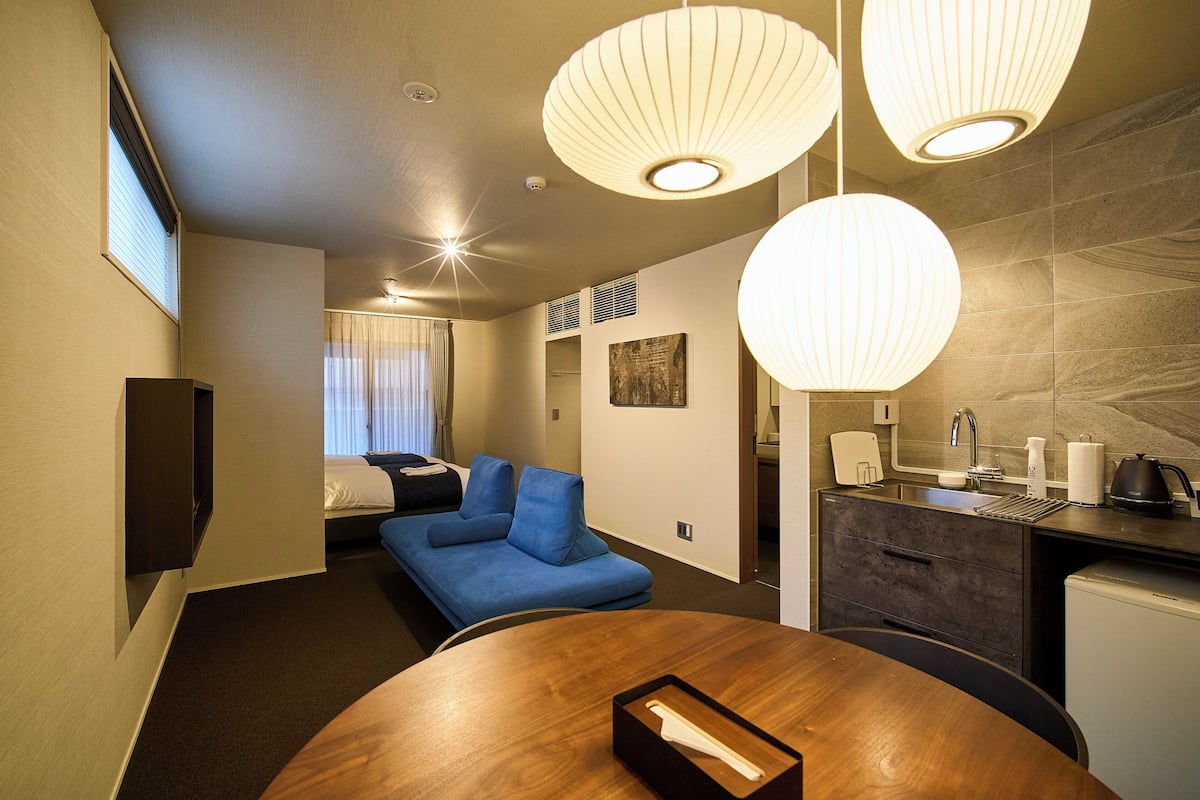 A modern hotel room with a blue sofa, a wooden dining table, and a kitchenette.