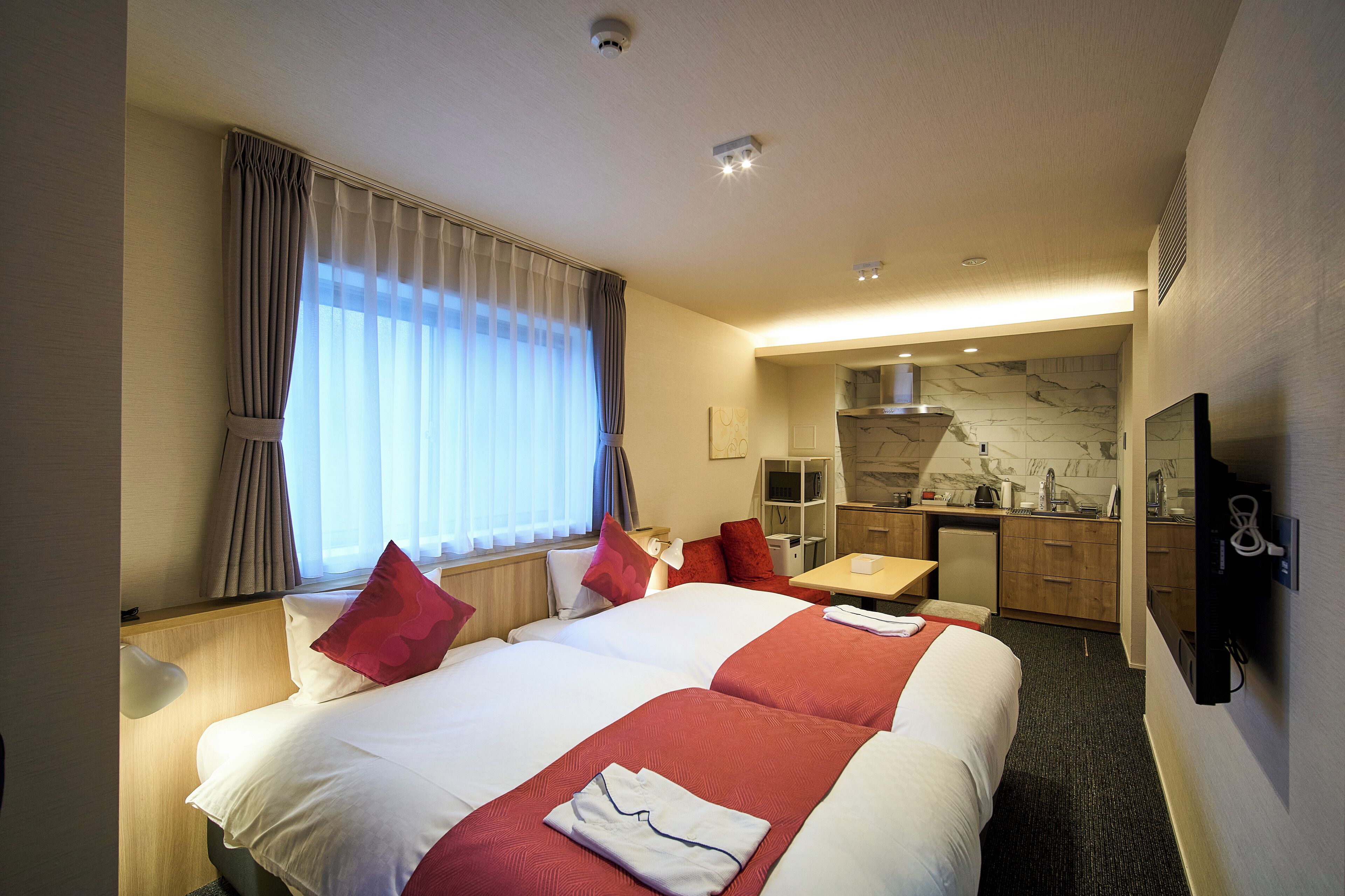 A hotel room with two beds, a desk, a kitchenette, and a TV.