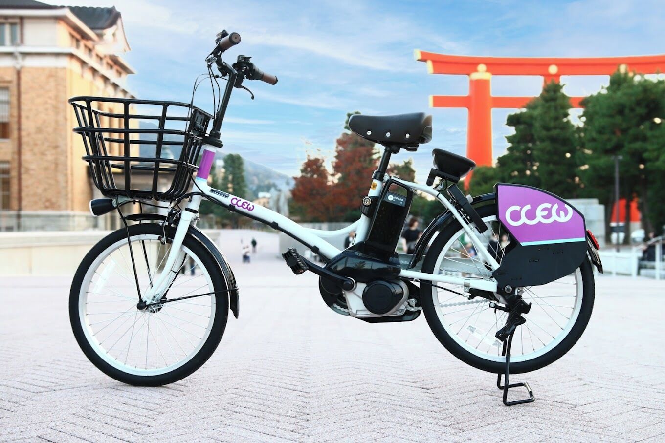 Una bicicleta eléctrica estacionada, con canasta y caja trasera, sobre una superficie pavimentada, con un torii rojo y árboles de fondo.