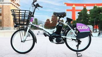 Una bicicleta eléctrica estacionada, con canasta y caja trasera, sobre una superficie pavimentada, con un torii rojo y árboles de fondo.