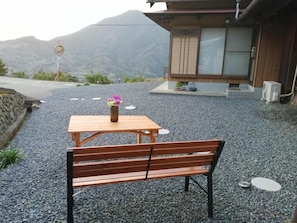 Un banc et une table en bois, surmontés d’un vase de fleurs, sont placés sur un sol de gravier. On aperçoit en arrière-plan une montagne et un bâtiment doté de portes coulissantes.