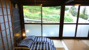 A traditional Japanese room with tatami flooring, a bed with striped bedding, a shoji screen, and a view of a hillside. - Kumano Kodo Nagano Guesthouse (Tanabe)