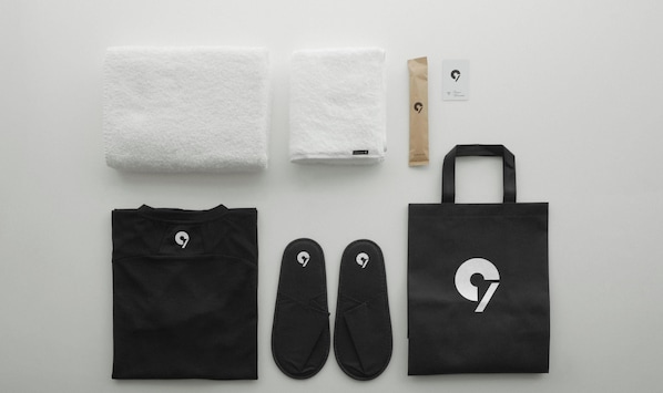 Una bolsa de tela negra, un par de zapatillas negras, una camiseta negra, dos toallas blancas, una bolsa de papel marrón y una tarjeta con un logotipo.