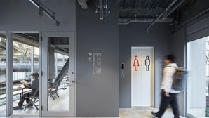 Un espacio interior con una escalera que baja a un nivel inferior, una puerta de entrada de vidrio y una pared con señalización direccional.