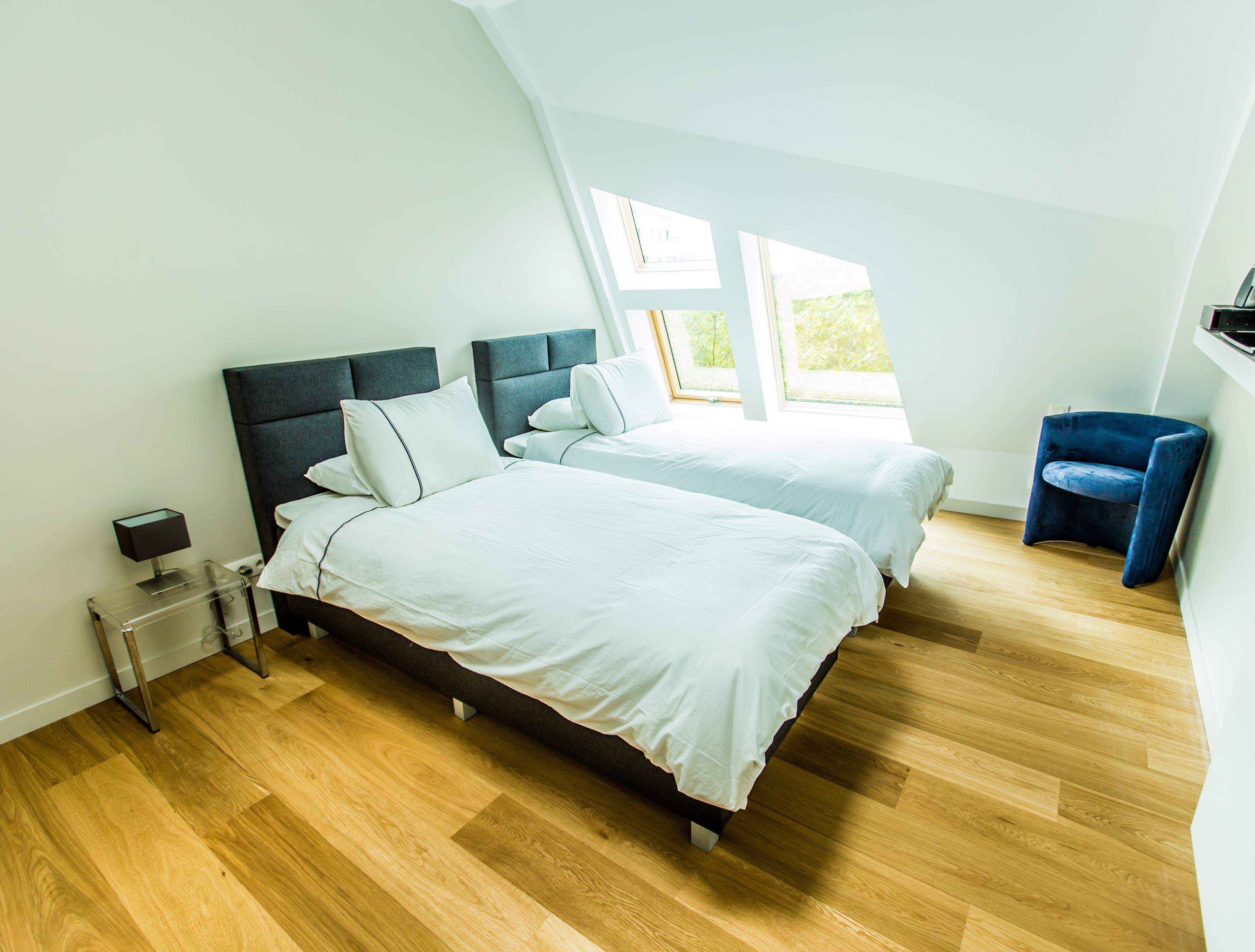 A modern bedroom with two beds, a nightstand, a chair, and large windows.