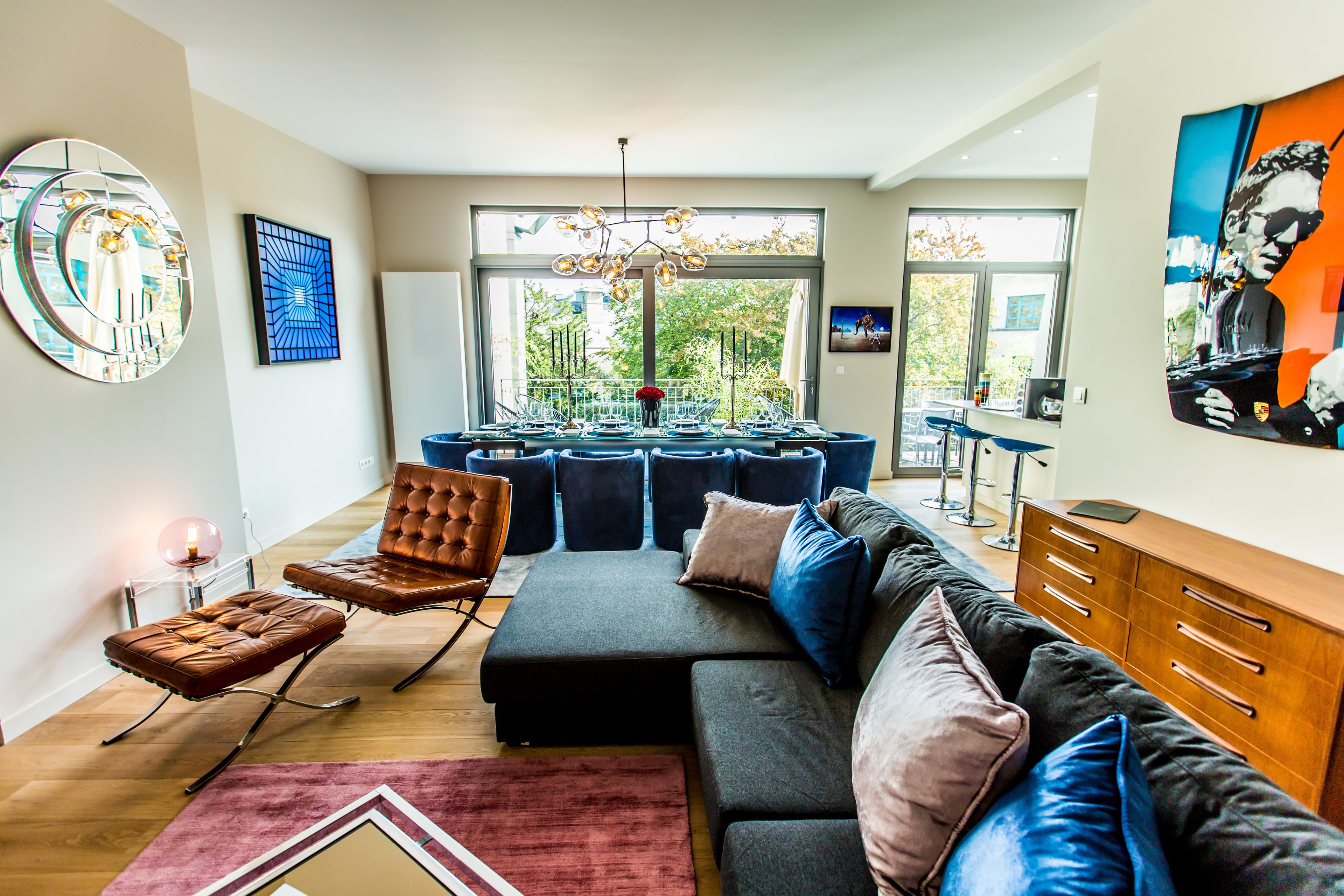 A modern living room with a sectional sofa, a dining area with round tables, and a large window offering a view of trees.