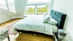 A bedroom with a large bed, a nightstand, a lamp, and a chair. There are multiple windows providing natural light and views of the outside.