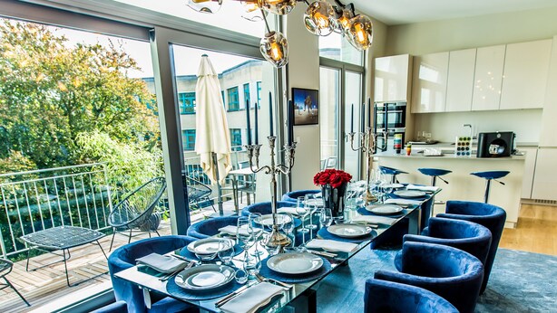 A modern dining area with a long table set for six, surrounded by blue chairs, a chandelier, and a view of the outdoors.