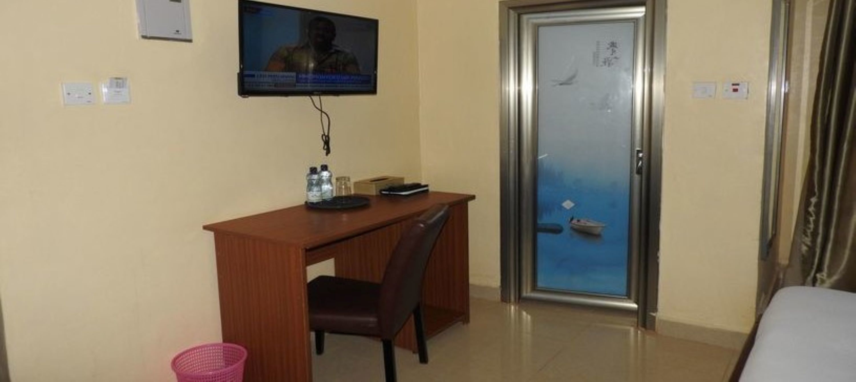 A hotel room with a wooden desk, a chair, a television mounted on the wall, a door with a view of the sea, and a small trash bin.