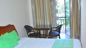 A hotel room with a bed, green pillows, a wooden headboard, a table with four chairs, and a balcony with curtains. - Omega Gardens Hotel (Karatina)