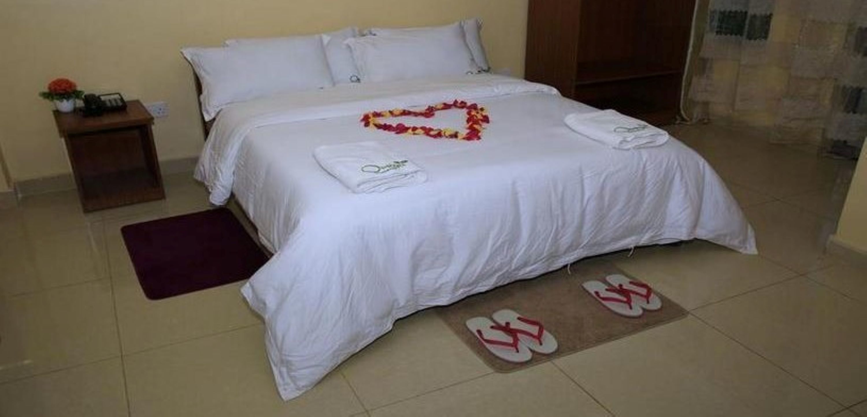 A neatly made bed with a heart-shaped flower arrangement, a folded towel, and a pair of slippers on the floor.