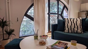 A cozy living room with a round white table, a teal sofa with patterned pillows, a potted plant, and a window with a view of trees. - Basco Central Apartment Little Bay (Belgrade)