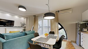 A modern living space with a sofa, dining table, chairs, a TV, and a kitchen area. - Basco Central Apartment Little Bay (Belgrade)