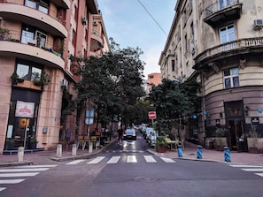 A street view with a crosswalk, parked cars, and buildings with balconies and signage. - Basco Central Apartment Little Bay (Belgrade)