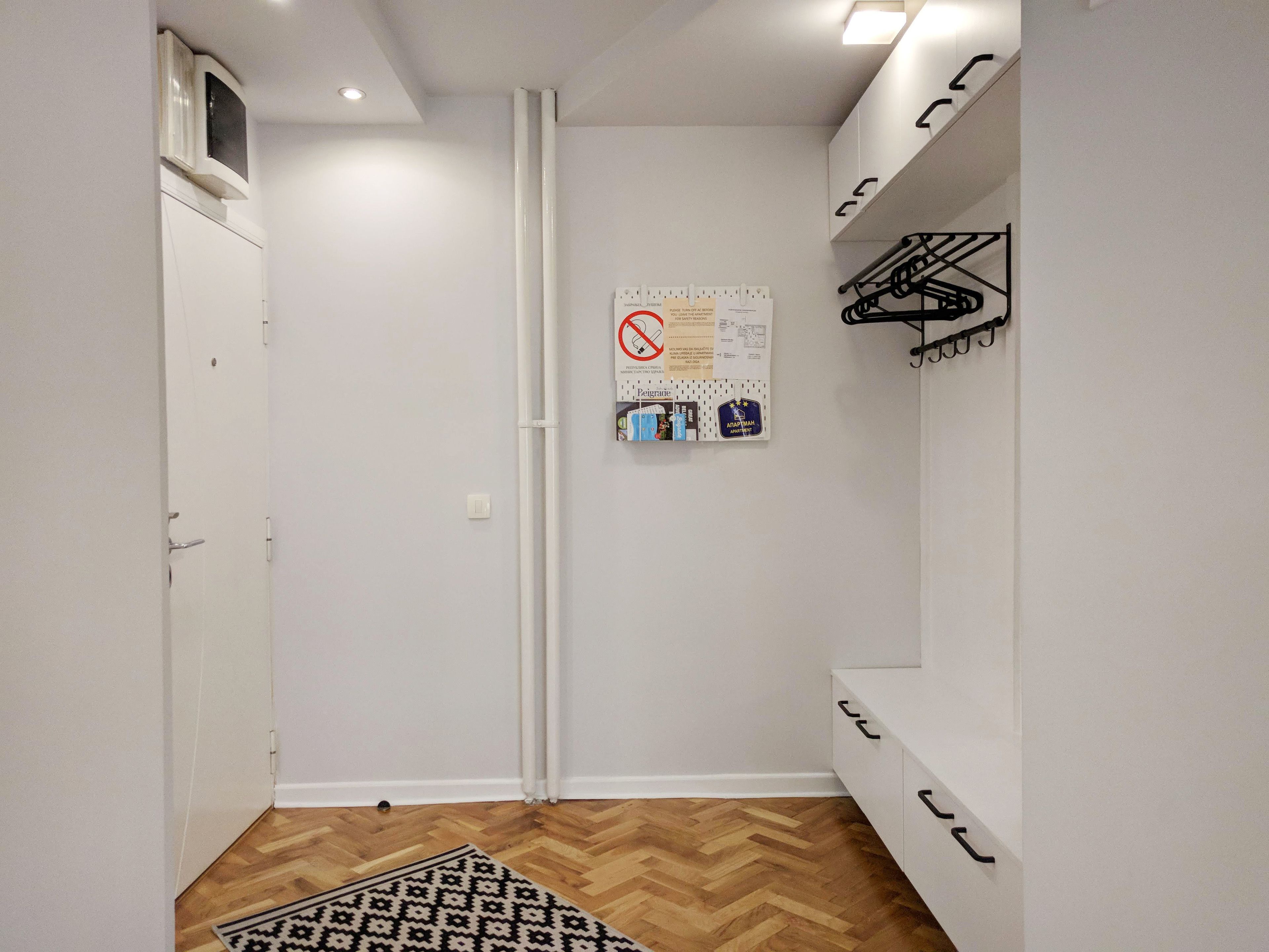A hallway with a white door, a bulletin board with notices, a black metal rack, and a patterned rug.