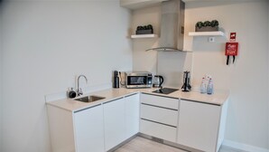 A modern kitchen with white cabinetry, a built-in microwave, a kettle, and a sink. There are two shelves above the counter, one with plants and the other with a red fire extinguisher. - Calamar Hotel (São Vicente)