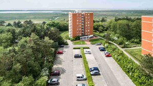 A multi-story building with a parking lot in front, surrounded by greenery and a distant view of a flat landscape.