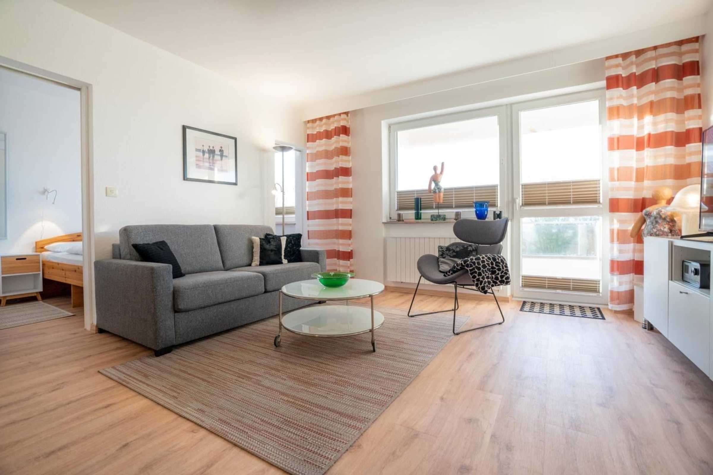 A modern living room with a grey sofa, a small round table with a green bowl, a black chair, and a window with striped curtains.