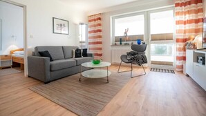 A modern living room with a grey sofa, a small round table with a green bowl, a black chair, and a window with striped curtains.