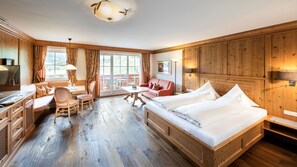 A hotel room with a wooden floor, a bed with white linens, a sofa, a desk with a chair, and a window with curtains.