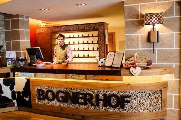 Una recepción con un cartel que dice 'BOGNERHOF', una exhibición de figuritas, una cesta con un objeto en forma de corazón y una lámpara fijada a la pared.