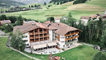 Ein großes, mehrstöckiges Gebäude im traditionellen alpinen Baustil, umgeben von üppigen grünen Feldern und Bergen im Hintergrund.