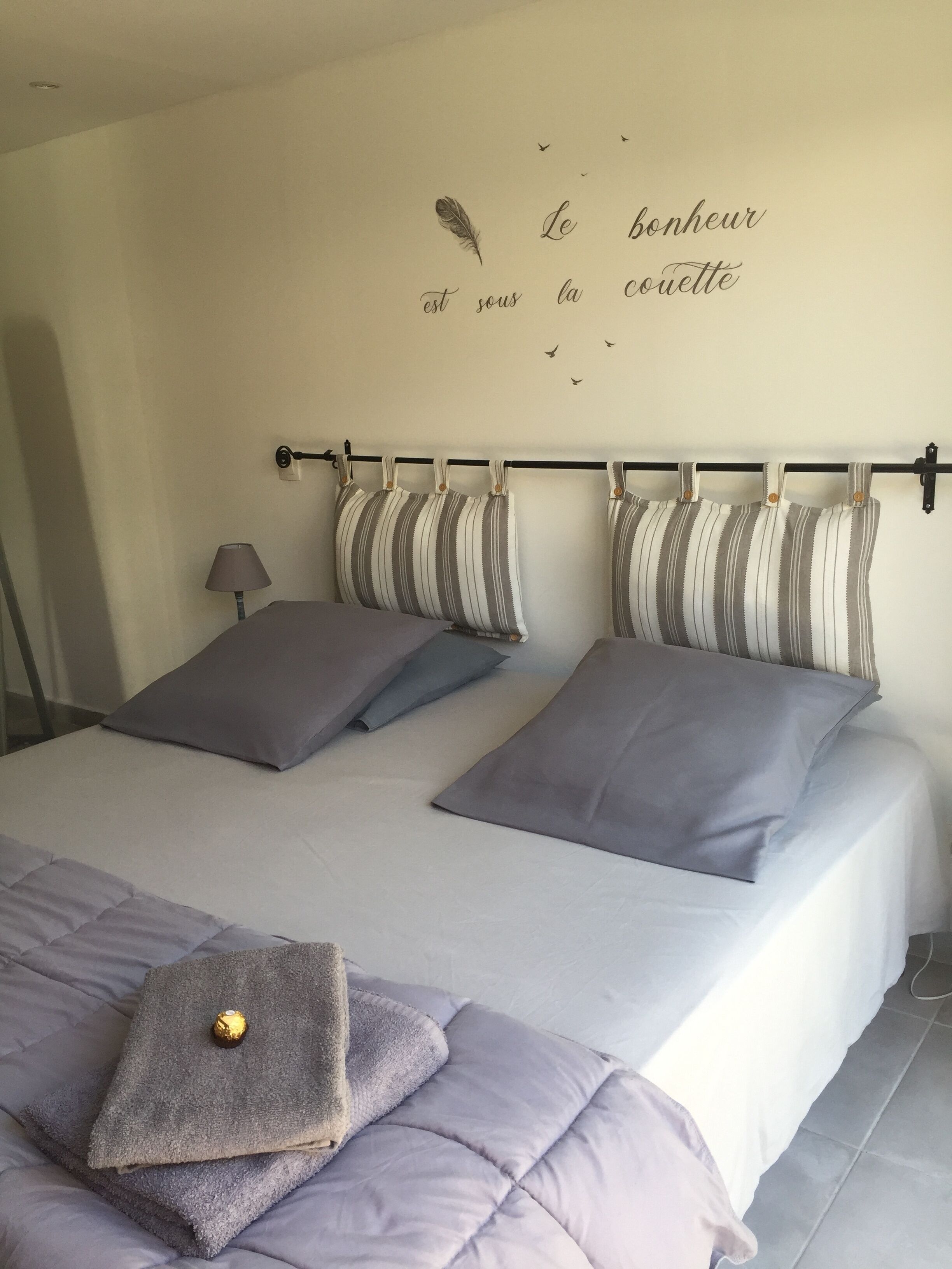 Een bed met grijze beddengoed, gestreepte kussens en een decoratieve muur met een Franse tekst.