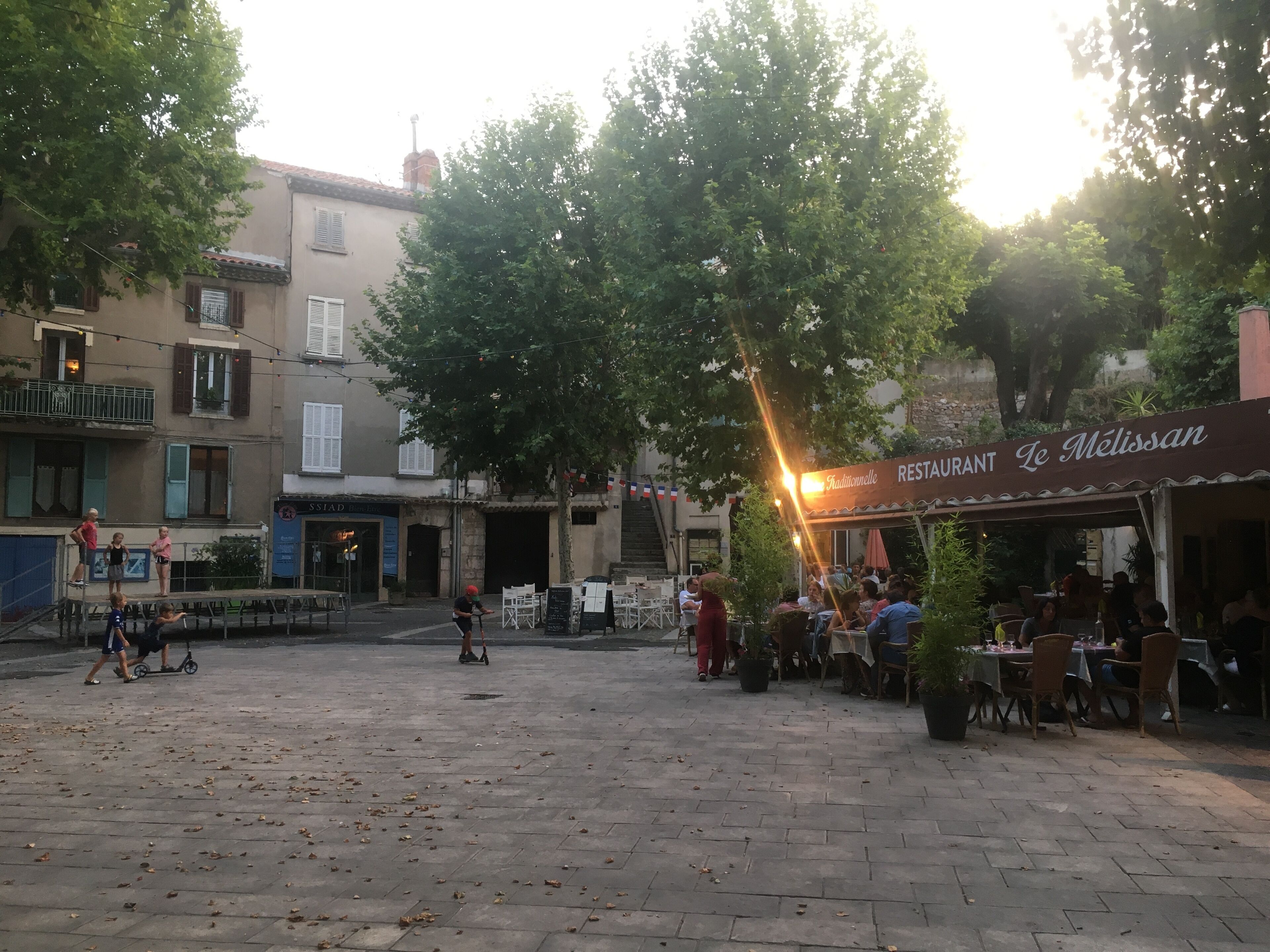An outdoor café area with tables and chairs, surrounded by buildings and trees.