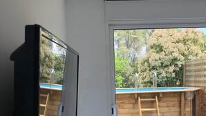 Une pièce dotée d’une porte-fenêtre coulissante donnant sur une terrasse en bois, accessible par une échelle, et qui offre un accès à une piscine.