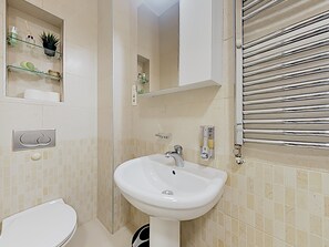 A bathroom with a white sink, a toilet, and a mirror above the sink. There are shelves with toiletries and a towel rack. - Basco Apartment Terazije Square (Belgrade)