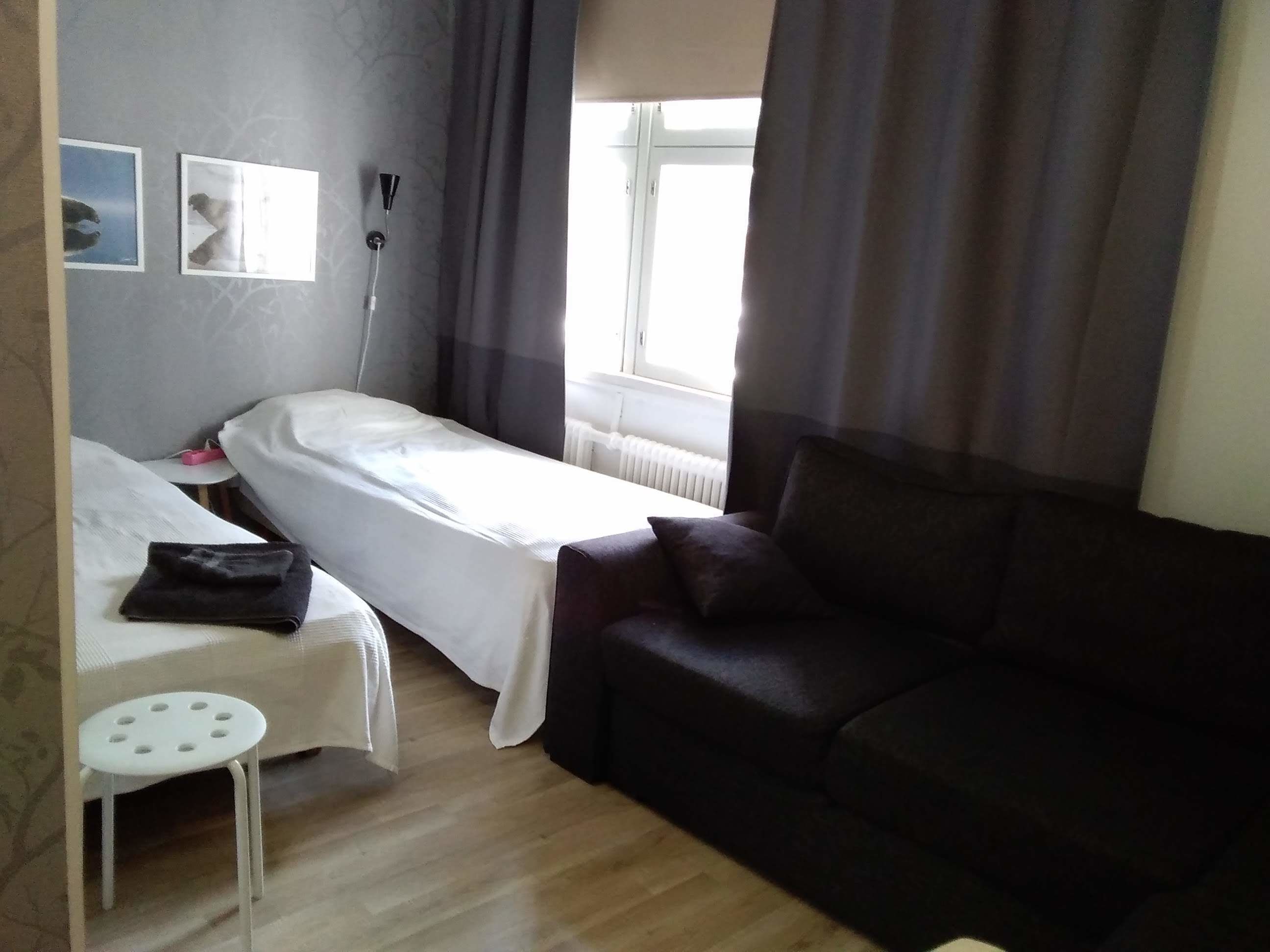 Una habitación pequeña y ordenada con una cama individual, un sofá oscuro, una mesita y una ventana con cortinas.