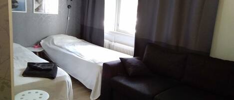 Een kleine, netjes ingerichte kamer met een enkel bed, een donkere bank, een tafeltje en een raam met gordijnen.