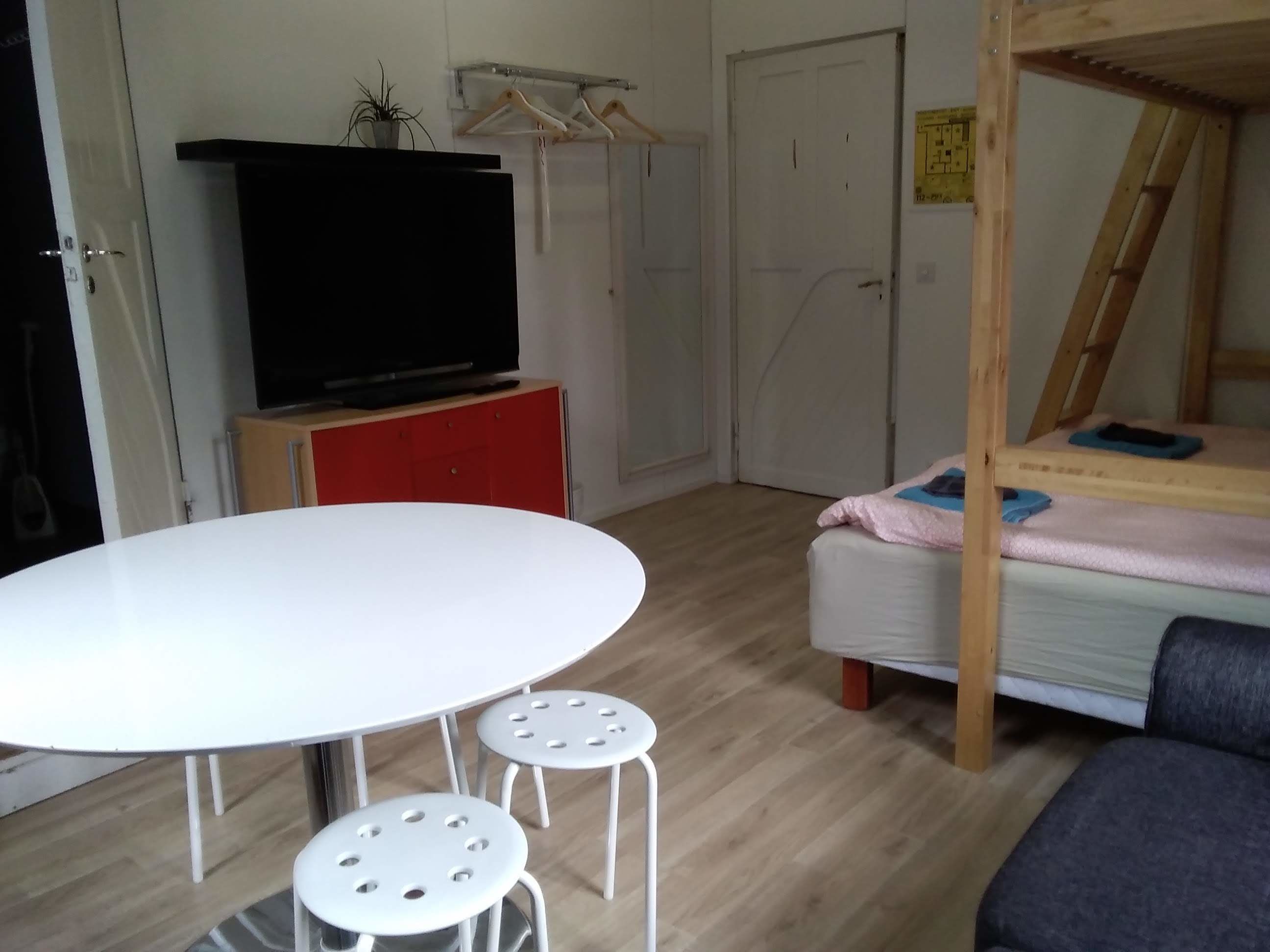 A room with a round white table, two stools, a bed with a wooden frame, a TV on a red cabinet, and a wardrobe with a mirror.