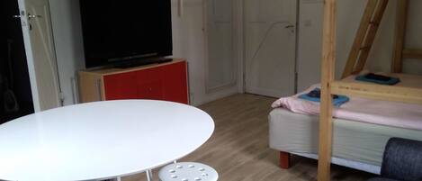 A room with a round white table, two stools, a bed with a wooden frame, a TV on a red cabinet, and a wardrobe with a mirror.