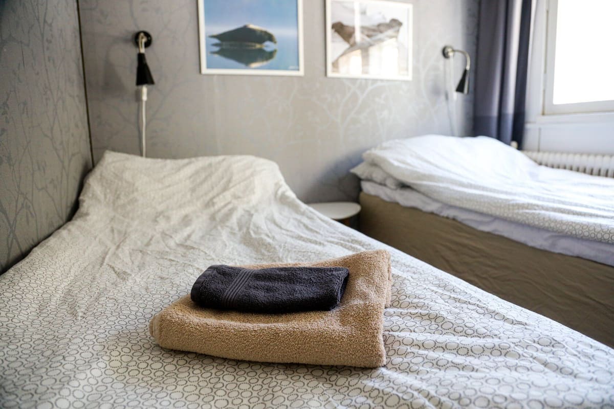 A neatly made bed with a folded towel and a pillow, a bedside table with a lamp, and a window with curtains.