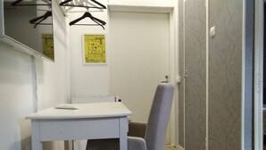 Een kleine kamer met een wit bureau, een grijze stoel, een spiegel en een deur met behang met een patroon.
