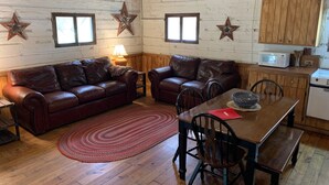 A cozy living room with a brown leather sofa, a wooden dining table with chairs, and a kitchen area with wooden cabinets and a microwave. - Rustic River Cabin under the Oaks and just a short walk to the river. (Uvalde)