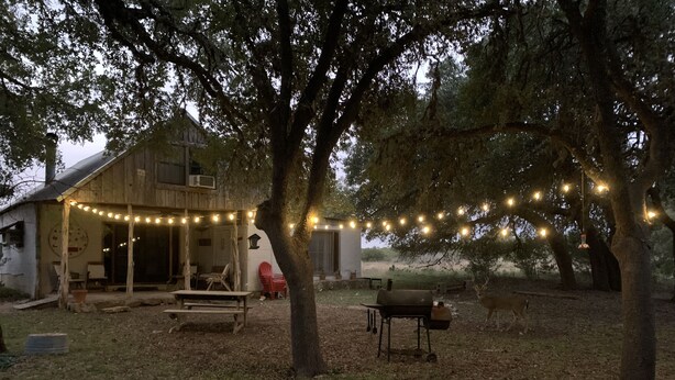 A rustic barn with string lights, a red chair, and a wooden table in the foreground. - Rustic River Cabin under the Oaks and just a short walk to the river. (Uvalde)