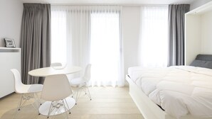 A modern hotel room with a bed, a round dining table with chairs, and a large window with curtains. - White Studio (Brussels)