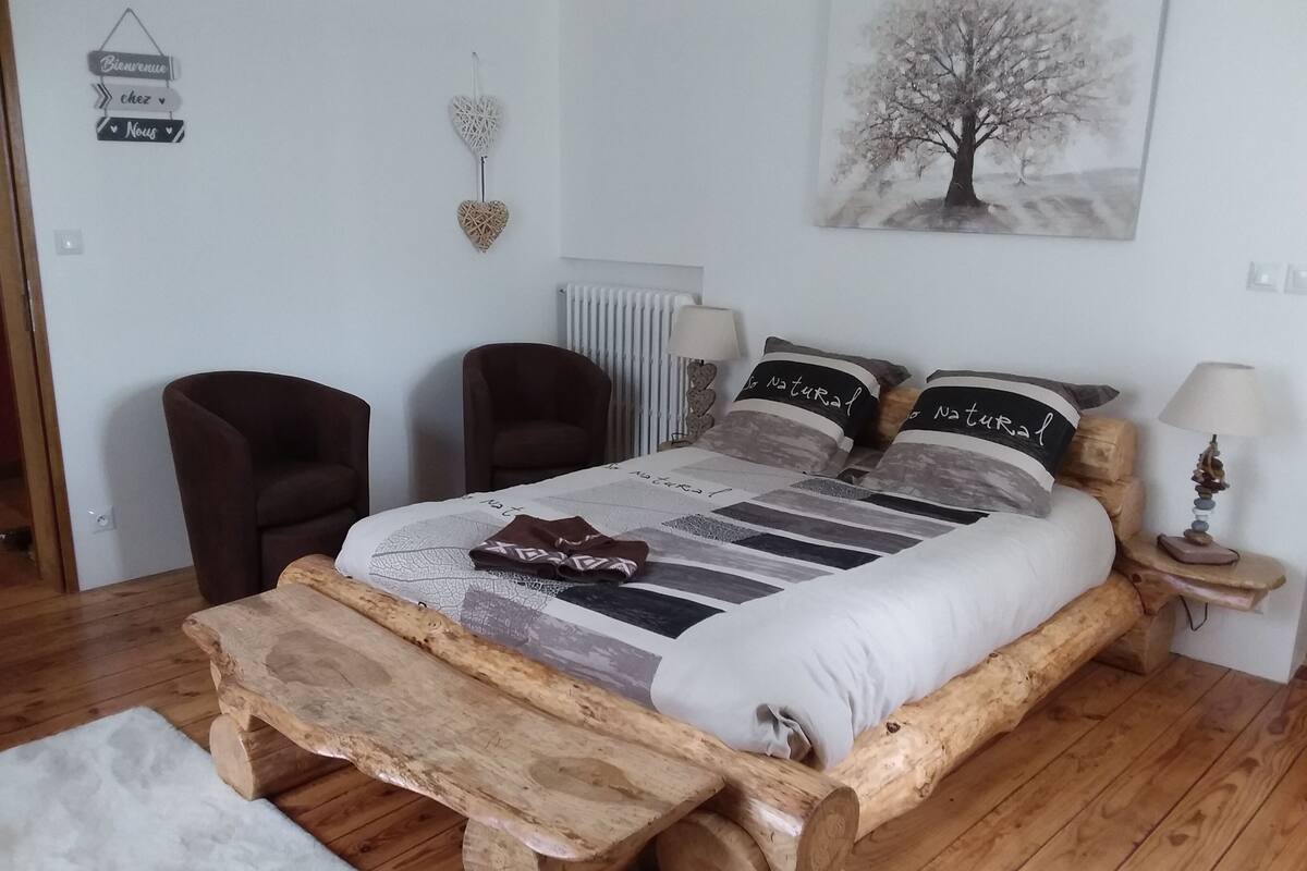 Een slaapkamer met een houten bed, twee stoelen, een nachtkastje met een lamp, een radiator en een bordje aan de muur.