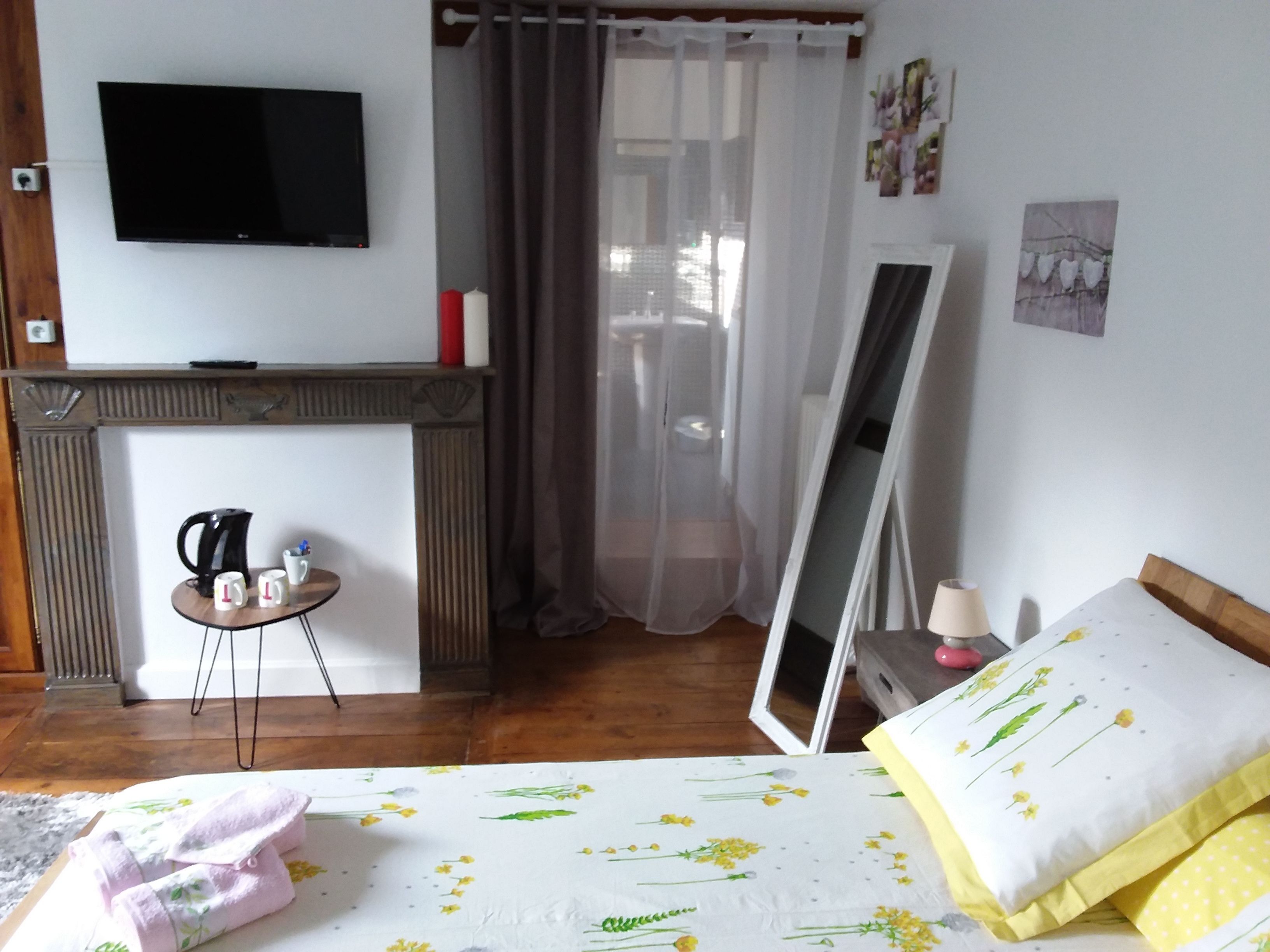 A bedroom with a bed, a bedside table with a lamp, a mirror, a fireplace, a TV, and a window with curtains.