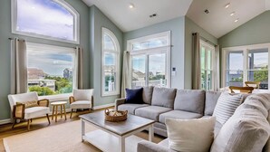 Un salon spacieux avec un grand canapé d’angle gris, deux fauteuils blancs, une table basse et une vue sur le quartier grâce à de grandes fenêtres.